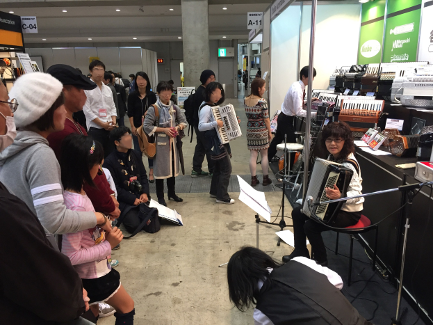 Eine Gruppe von Menschen, die Musikinstrumente um einen Tisch spielen, mit einer Frau in der Mitte, die eine Akkordeon spielt, einem Tisch mit Gegenständen rechts daneben und Tafeln mit Text, einer Säule und Deckenlampen im Hintergrund auf einem japanischen Musikfestival.