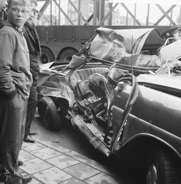 Schwarzes und weißes Bild eines verbeulten Autos am Straßenrand mit Menschen in der Nähe, einem Lastwagen und einer Brücke im Hintergrund.