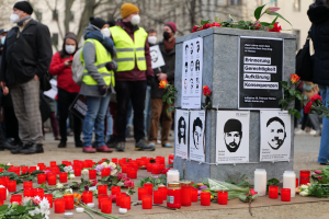 Eine Gruppe von Menschen steht ernst vor einem mit Kerzen, Blumen und Plakaten geschmückten Denkmal, umgeben von Bäumen und Gebäuden mit Fenstern im Hintergrund, und gedenkt einer Tragödie in Berlin, Deutschland.