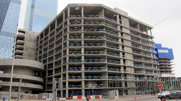 Großes Bauwerk im Bau in einer städtischen Umgebung mit Wolkenkratzern, Fahrzeugen, Fußgängern, Verkehrsinfrastruktur und Kränen vor einem klaren Himmel.