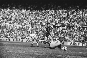 Schwarzes und wei├čes Foto von M├Ąnnern, die auf einem Feld Fussball spielen, mit Zuschauern im Hintergrund und einem Banner mit der Aufschrift "Bundesliga 1978-79 Ajax 1-1 Hannover 96."