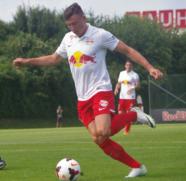 Ein professioneller Fußballspieler schießt einen Ball auf einem Rasenfeld mit Bäumen und einem klaren blauen Himmel im Hintergrund, in der Nähe eines Schildes mit der Aufschrift "RB Leipzig."