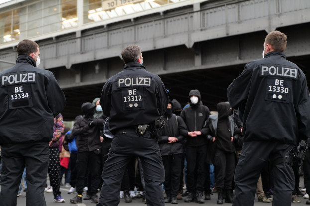 Polizeibeamte in schwarzen Uniformen und Masken vor einer Menge bei einer Demonstration, mit einer Brücke und einem Gebäude im Hintergrund.