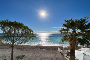 Ein sonniger Strand mit Palmen, Schirmen, üppiger Vegetation und einem strahlend blauen Himmel an der französischen Riviera.