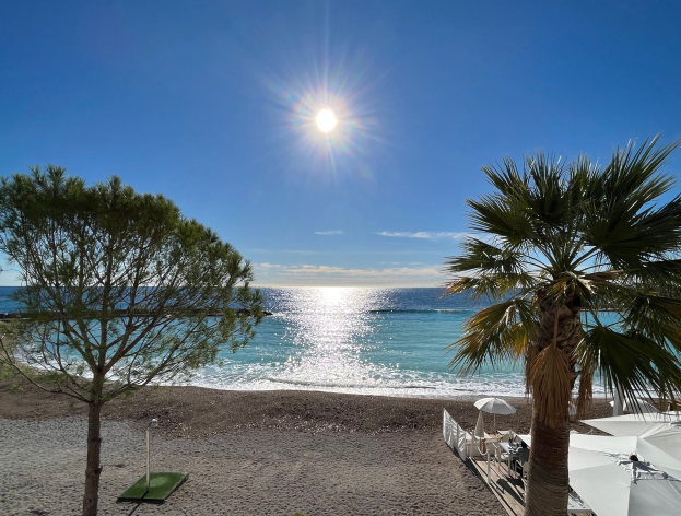 Ein sonniger Strand mit Palmen, Schirmen, üppiger Vegetation und einem strahlend blauen Himmel an der französischen Riviera.