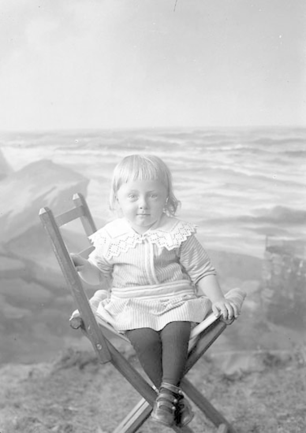 Ein kleines Mädchen sitzt in einem Stuhl am Strand, mit dem Meer und dem Himmel im Hintergrund, auf einem alten Schwarz-Weiß-Foto.