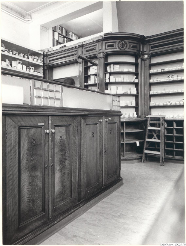 Ein Schwarz-Wei├č-Foto eines Apothekeninneren mit einer Theke, Sch├Ąben, einer Leiter und Regalen, die mit verschiedenen Gegenst├Ąnden gef├╝llt sind, mit Text ├╝ber dem Bild.