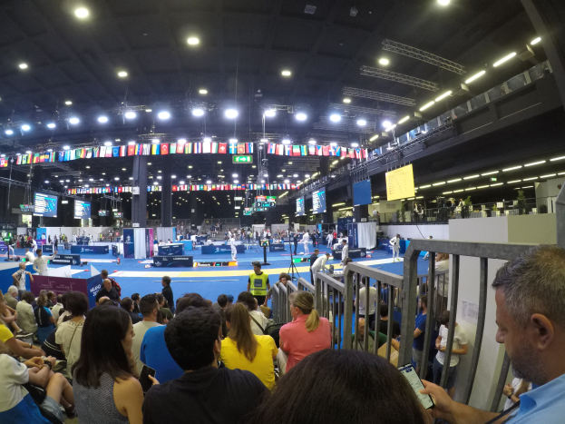Eine große Menge an Sitz- und Stehplätzen in einer Indoor-Sportarena mit Bildschirmen, Fahnen und Deckenbeleuchtung im Hintergrund.