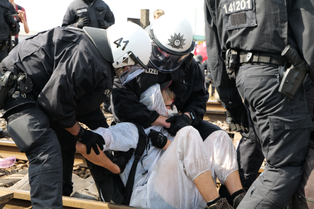 Eine Gruppe von Polizisten in Einsatzausrüstung verhaftet einen Mann in einem weißem Kleid auf einem Gleis, mit anderen im Hintergrund sichtbar.