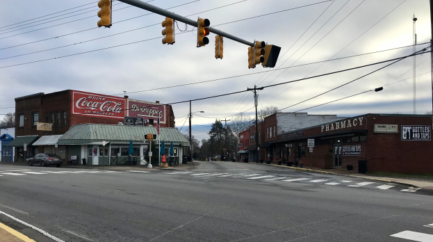Eine Stadtstraßenkreuzung mit Verkehrsampel, Fahrzeugen, Gebäuden mit Namensschildern, Strommasten mit Kabeln, Bäumen und einem bewölkten Himmel.
