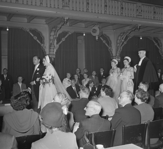 Ein Schwarz-Weiß-Foto einer Hochzeitszeremonie in einem großen Raum mit vielen Sitzenden und Stehenden, einige halten Blumensträuße, und Tische mit Tassen und anderen Gegenständen davor, mit Vorhängen und Deckenlampen im Hintergrund.