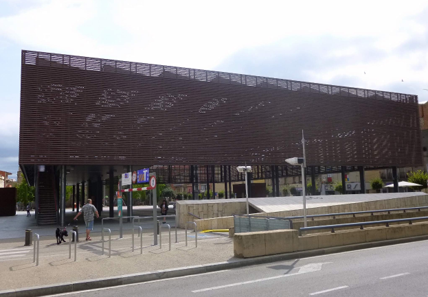 Großes modernes Gebäude mit zahlreichen Fenstern, das als neuer Eingang zur City dient, umgeben von städtischen Elementen wie Straßenlaternen, Fahrzeugen, Fußgängern und Bäumen unter einem bewölkten Himmel.
