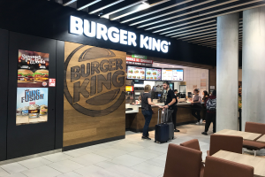 Ein Burger King Restaurant am Flughafen mit Menschen mit Gepäckwagen, Tischen, Stühlen, einer Säule, einem Schild, einem Anzeigebildschirm und Deckenleuchten.