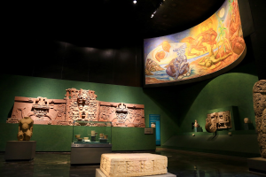 Ein Museumsraum mit verschiedenen Artefakten, darunter Skulpturen, Tischobjekte, eine Texttafel, eine Wandmalerei und Deckenleuchten im Anthropologischen Museum von Mexiko-Stadt.