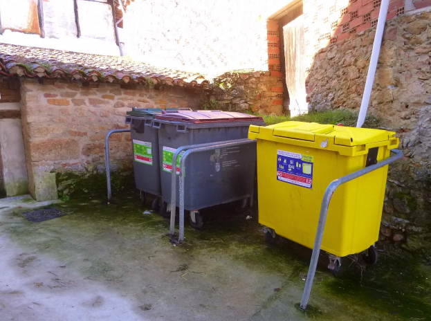 Zwei gelbe und graue Recyclingcontainer auf Rädern nebeneinander vor einem Gebäude mit einer Wand und einem Fenster im Hintergrund.