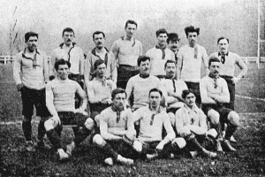 Ein Schwarz-Weiß-Foto einer Fußballmannschaft aus den 1920er Jahren, mit einigen stehenden und anderen auf dem Boden sitzenden Spielern, vor einem Hintergrund aus Pfählen, Bäumen und einem klaren Himmel.