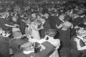 Ein Schwarz-Weiß-Foto einer Gruppe von Menschen, die auf einer Tanzfläche tanzen, einige halten Kameras, mit Treppen und Lichtern im Hintergrund.