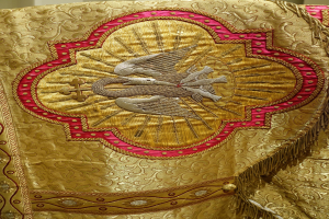 Nahaufnahme eines Priestergewandes mit aufwendiger goldener und roter Stickerei vor einer Wand im Hintergrund.
