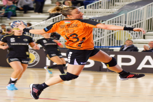 Eine Frau in einem orangen T-Shirt und schwarzen Shorts hält einen Handball, mit Zuschauern und einem Banner "Krp Slezak - Handball-Weltmeisterschaft 2019" im Hintergrund.