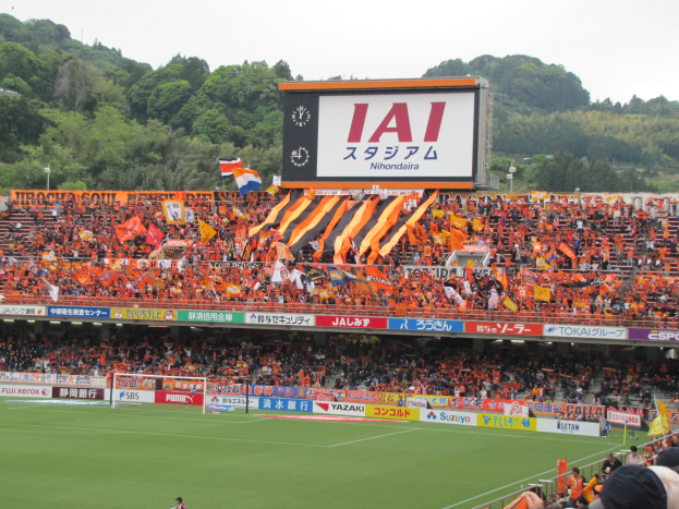 Fußballspiel in einem Stadion mit Zuschauern, saftigem Grün, Tor, Bannern, Fahnen, Großbildschirm, Bäumen und wolkenlosem Himmel.