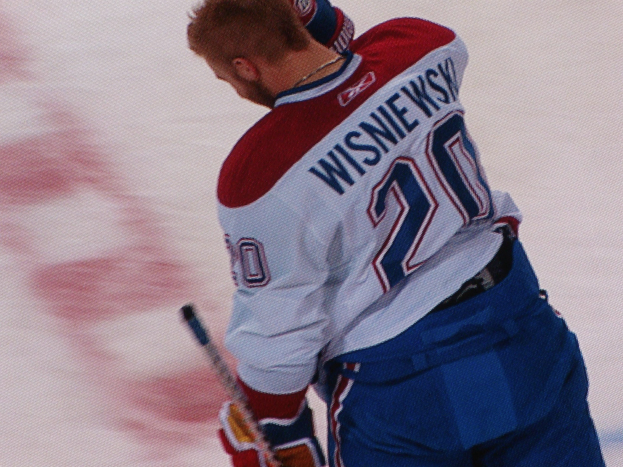 Ein konzentrierter Profi-Hockeyspieler im "Wisniewski"-Trikot steht auf dem Eis und hält einen Hockey-Schläger, entschlossen.