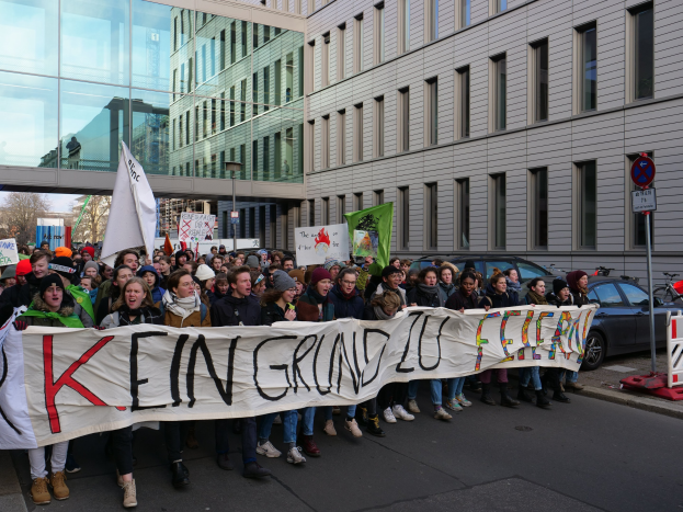 Protestierende auf einer Straße halten ein Banner mit der Aufschrift "Kein Grind Lo Feuer" gegen deutsche Sparmaßnahmen, mit Gebäuden, Bäumen und Fahrzeugen im Hintergrund unter einem klaren blauen Himmel.