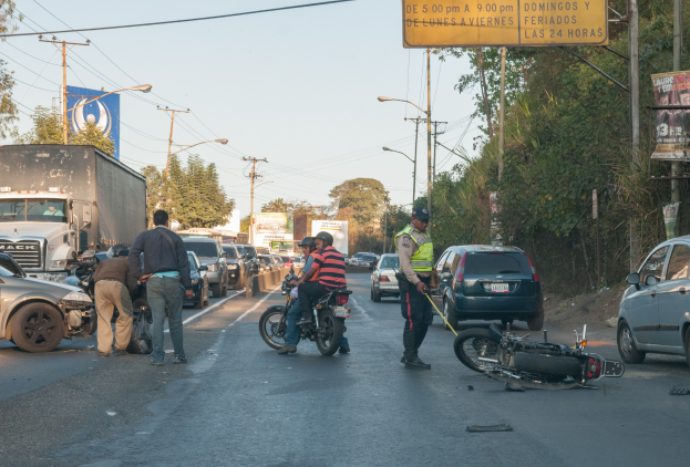 Gruppe von Menschen um ein verunglücktes Motorrad am Straßenrand mit mehreren Fahrzeugen, darunter ein Lastwagen, im Hintergrund und Bäumen, Pfosten, Lichtern, Schildern und Himmel.