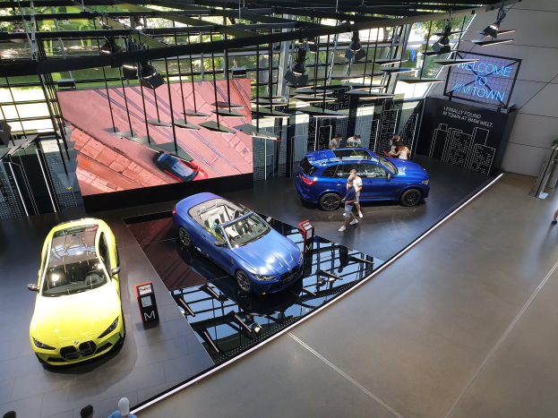 Eine Gruppe von Menschen im BMW Museum in München, Deutschland, mit Autos auf der linken Seite und einem Schild mit der Aufschrift "BMW Museum München" auf der rechten Seite, mit Blick auf eine Außenansicht von Bäumen, Gebäuden und Himmel durch Glasfenster.