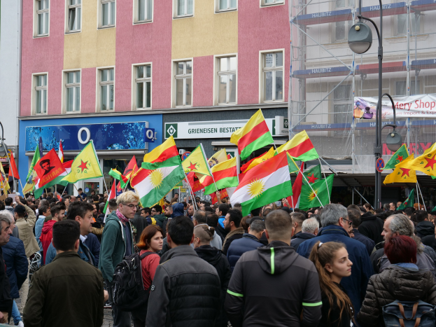 Eine große Gruppe von Menschen geht eine Straße entlang und hält kurdische Flaggen in den Händen.