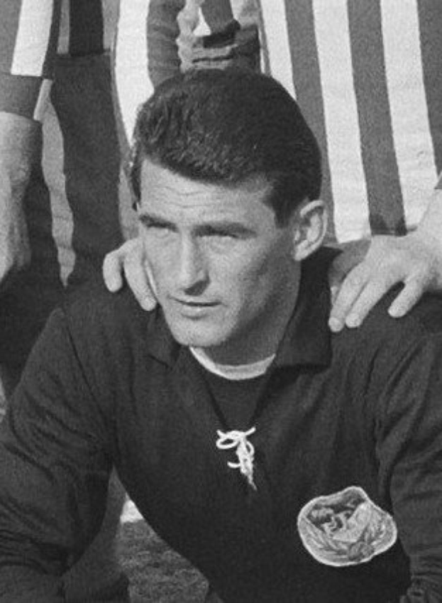 Schwarzes und weißes Foto eines Mannes, der auf einem grasbewachsenen Fussballfeld sitzt und ein T-Shirt und eine Kette trägt, mit zwei Personen hinter ihm.