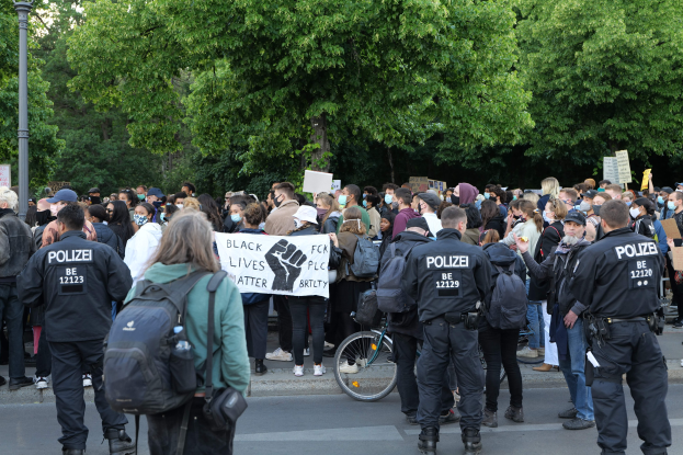 Große Gruppe von Menschen an einer Straße mit Schildern, einige tragen Mützen und Taschen, mit einem Fahrrad im Vordergrund und Bäumen und einem Pfahl im Hintergrund.