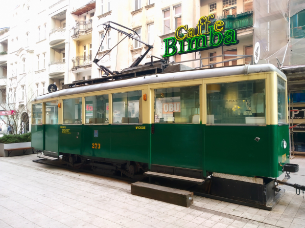 Ein grüner und gelber Tram fährt auf einer städtischen Straße mit Gebäuden, die Fenster, Geländer, Balkone und Namensschilder aufweisen, sowie Bäumen und Pflanzen im Hintergrund unter einem klaren blauen Himmel.