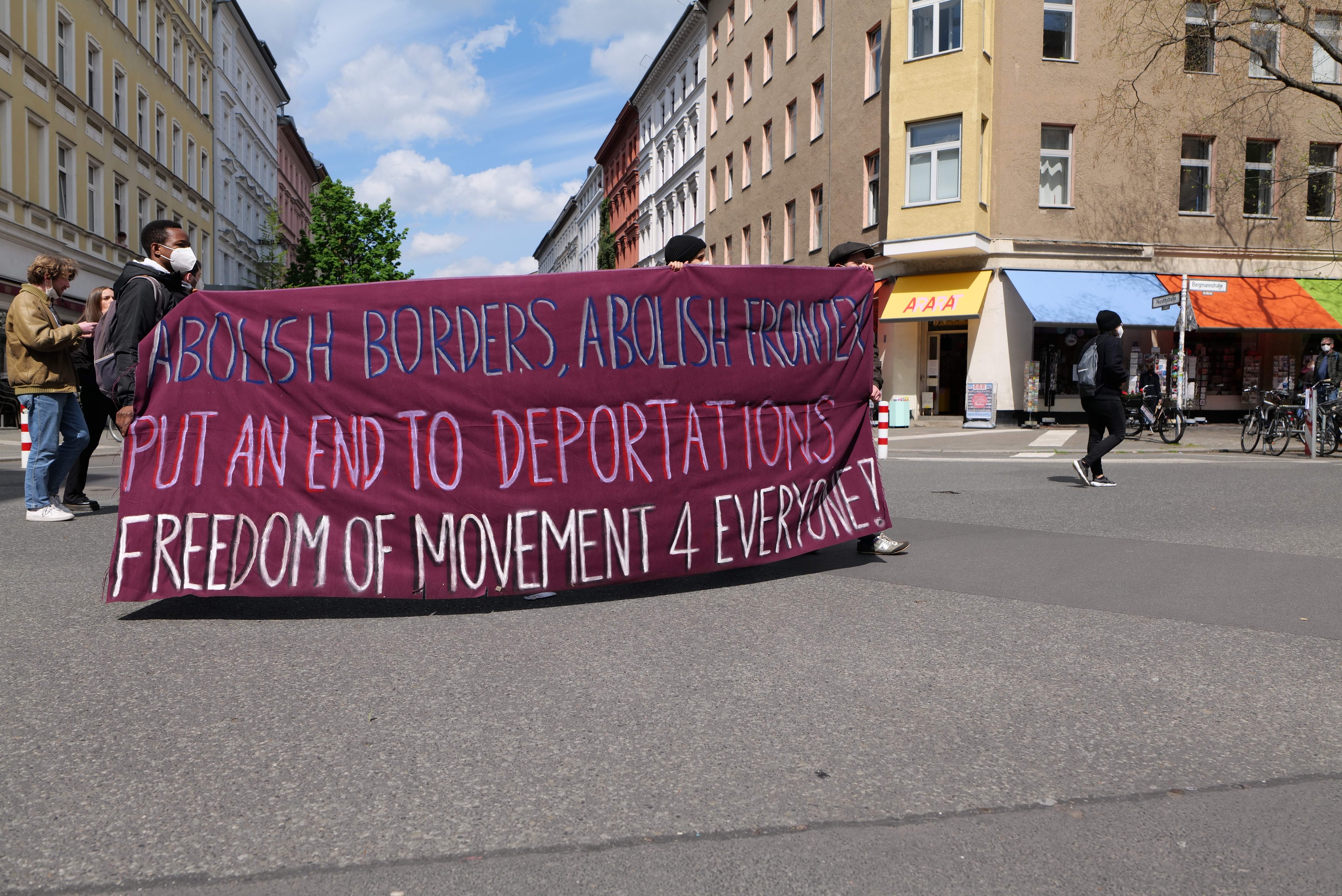 Eine Gruppe von Menschen marschiert auf einer Straße mit einem Banner, auf dem "Abolish Borders, Abolish Frontiers, Put an End to Deportations, Freedom of Movement 4 Everyone" steht, mit Gebäuden, Bäumen, Fahrrádern und bewölktem Himmel im Hintergrund.