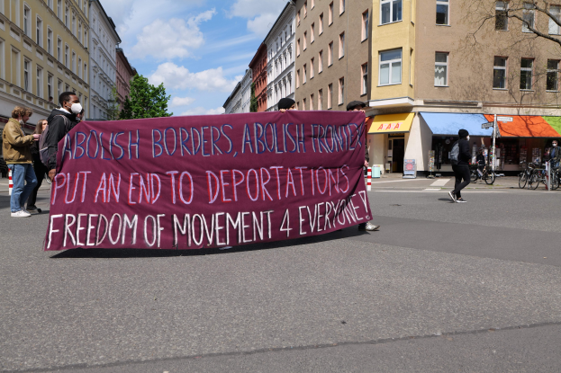 Eine Gruppe von Menschen marschiert auf einer Straße mit einem Banner, auf dem "Abolish Borders, Abolish Frontiers, Put an End to Deportations, Freedom of Movement 4 Everyone" steht, mit Gebäuden, Bäumen, Fahrrádern und bewölktem Himmel im Hintergrund.