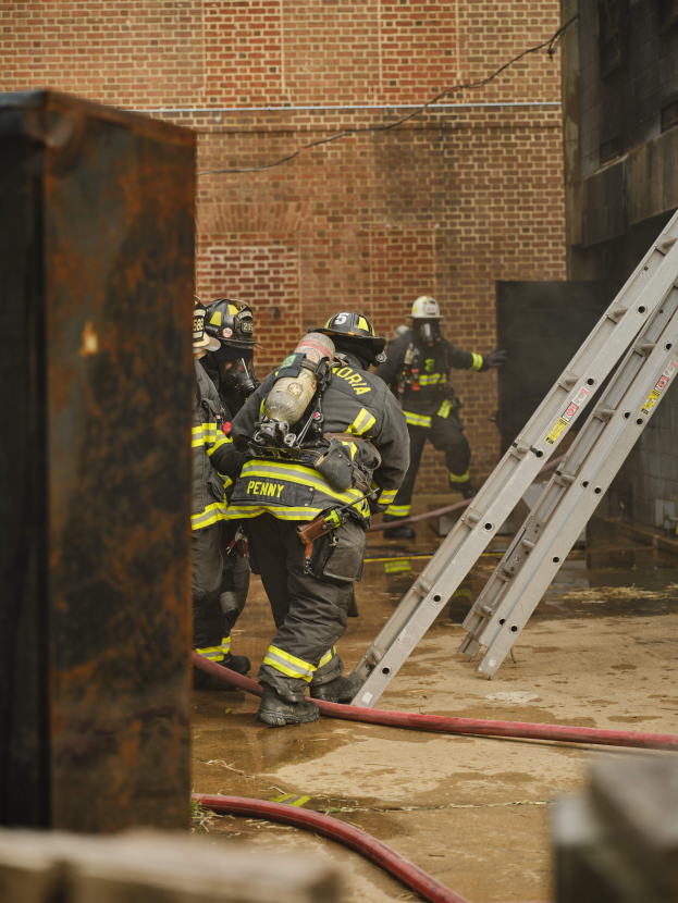 Feuerwehrleute in Helmen und Handschuhen arbeiten daran, ein Gebäude Feuer zu löschen, mit einer Leiter auf der rechten Seite und einem Metallobjekt auf der linken Seite, vor einem Ziegelmauer-Hintergrund.