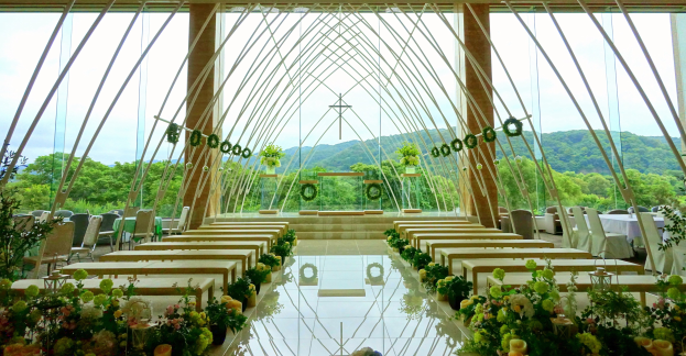 Eine Hochzeitszeremonie in einem großen, geschmückten Saal mit halbrunder Sitzordnung, sichtbarem Boden und Außenansicht von Bäumen, Hügeln und einem klaren blauen Himmel.