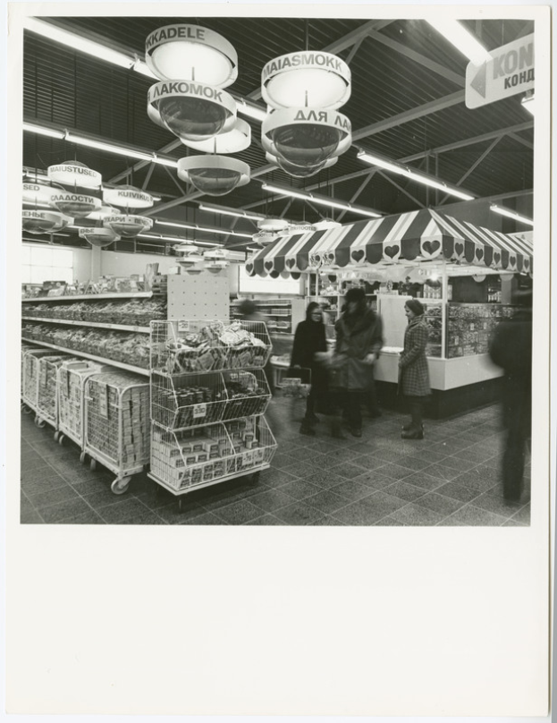 Ein Schwarz-Weiß-Foto von Menschen beim Einkaufen in einem Supermarkt mit vollen Einkaufswagen, Deckenleuchten und Wandschildern.