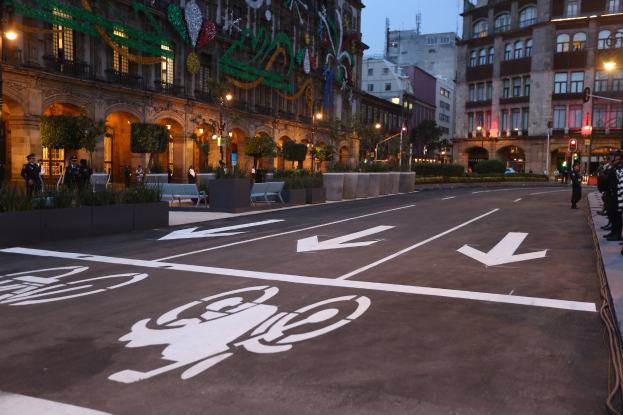 Stadtstraße mit einem Fahrradweg, umgeben von Gebäuden, Straßenmöbeln, Fußgängern auf dem Gehweg und Grünflächen unter einem klaren blauen Himmel.