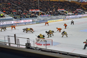 Eine Gruppe von Menschen, die Hockey auf einer Indoor-Eisbahn spielen, umgeben von einem Zaun, mit Zuschauern im Hintergrund und beleuchteten Bannern darüber.