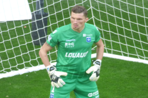 Ein Torwart in grüner Uniform steht vor einem Fußballtor, trägt Handschuhe auf einem grünen Rasen.