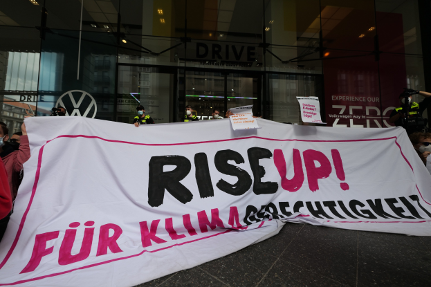 Eine Gruppe von Menschen mit Masken, die eine 'Aufstehen' -Plakette und Schilder vor einem Glasgebäude halten, wahrscheinlich als Protest gegen eine Entscheidung der deutschen Regierung bezüglich der Frauenrechte.