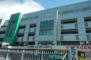 Ein großes Krankenhausgebäude mit einer 'Notaufnahme'-Schilder, umgeben von einem Metallzaun, Pflanzen, Texttafeln, Fahrzeugen und Klimaanlagen unter einem bewölkten Himmel.