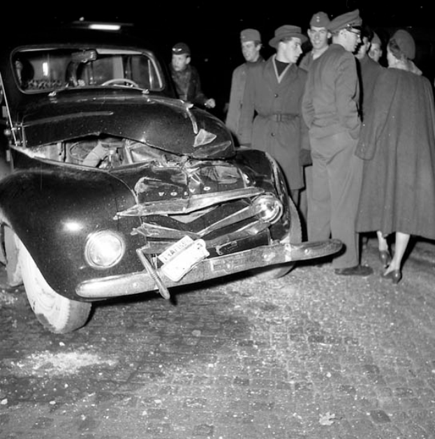 Eine schwarz-weiße Aufnahme eines beschädigten Autos am Straßenrand, umgeben von einer Gruppe besorgter Menschen.