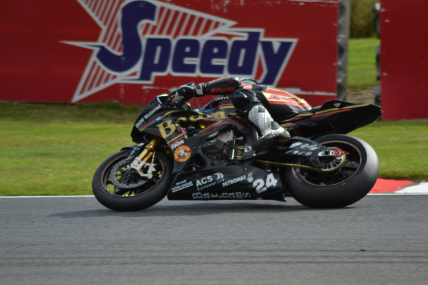 Eine Person in Helm und Schutzkleidung fährt ein Motorrad auf einer Rennstrecke, die von Gras umgeben ist, mit einer Tafel im Hintergrund, auf der Text zu sehen ist.