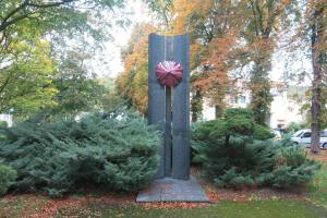 Große Metallskulptur in einem Park mit Bäumen, Pflanzen, Gras und welken Blättern umgeben von Gebäuden und Fahrzeugen unter einem klaren blauen Himmel, Teil der Kunst der Stadt Berlin.