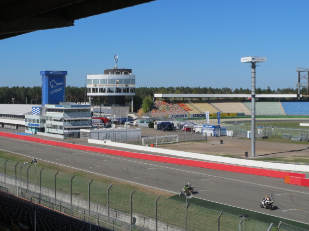 Gruppe von Motorradfahrern auf einer Rennstrecke, umgeben von einem Metallzaun, Gras, Fahrzeugen, einem Gebäude, einem Schild, Pfosten, einem Stadion, Deckenleuchten, Bäumen und einem bewölkten Himmel.