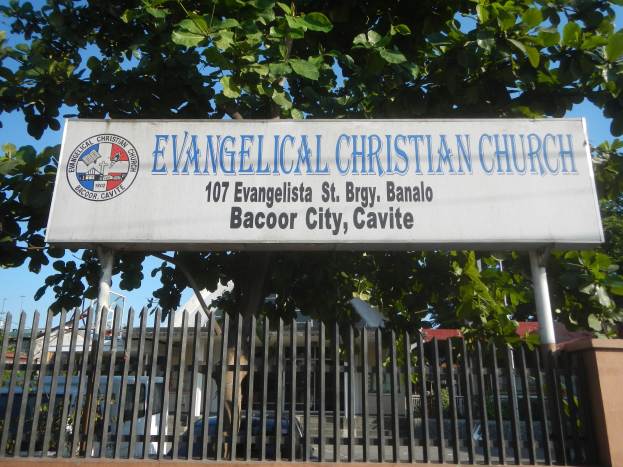 Evangelische Kirche in Bacoor City, Cavite, mit einem Schild, einem Metallzaun, einer Wand, Bäumen, geparkten Fahrzeugen, benachbarten Gebäuden, Strommasten und einem bewölkten Himmel.