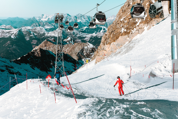 Gruppe von Menschen, die eine schneebedeckte Abfahrt mit einem Seilbahnzug im Hintergrund Ski fahren; eine Person in roter Kleidung mit Skistöcken im Vordergrund vor einer Bergkette und einem klaren blauen Himmel;