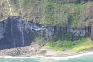 Ein Hubschrauber fliegt über das Meer nahe einer Klippe, mit Wasser darunter, Felsen und Gras am Boden und Bäume auf dem Berg im Hintergrund.