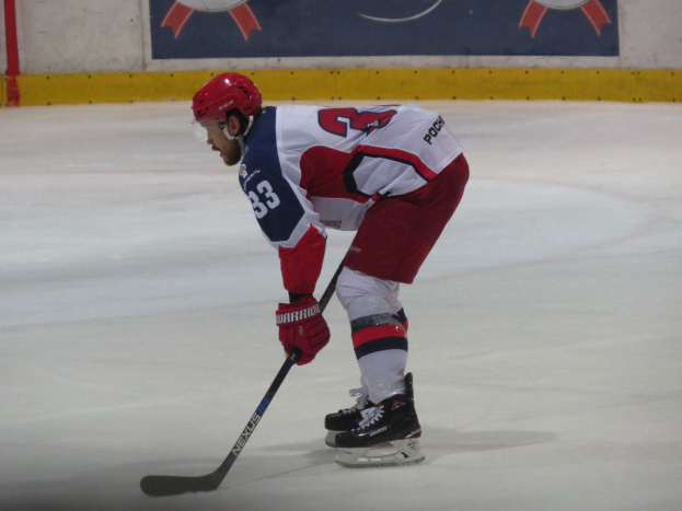 Ein Hockey-Spieler in voller Ausrüstung fährt auf dem Eis, hält einen Stock in der Hand und hat ein Banner mit Text und Logos im Hintergrund.